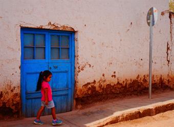 Ein weißes Haus mit einer großen leutend blauen Tür, davor ein rostfarbener Gehweg. Direkt vor der Tür geht ein kleines Mädchen mit schwarzen Haaren und einem pinken Tshirt.